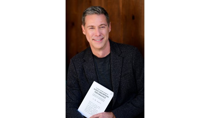 photo of Roy Samuelson holding his book 'Audio Description Performance'.  He is wearing a dark sport jacket with a dark crew-neck shirt and is in front of a dark wood wall.